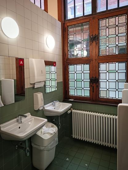 Two white ceramic sinks with modern chrome taps are mounted on a green-tiled wall in the men’s restroom of the Plantin-Moretus Museum in Antwerp. Above them are mirrors and round wall lights, as well as dispensers for soap and paper towels. A waste bin below holds used paper towels. Behind the sinks is a prominent wooden-framed window, original to the 16th-century structure. Its ornate leaded-glass design, featuring a pattern of diamond panes, is partially frosted for privacy. Through the clear sections, the viewer can see the red brick architecture and mullioned windows of the museum’s historic courtyard. A white radiator sits below the window, blending the historic ambiance with modern functionality.