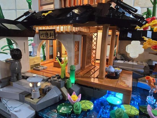 Eine "Bambushütte" aus Klemmbausteinen. Zu sehen ist das untere Stockwerk in Nahaufnahme mit Sicht in das "Wohnzimmer" und auf die Veranda, auf der ein Teeservive steht. Im Wasser sind Seerosen angedeutet und ein Koi springt aus dem Wasser.
