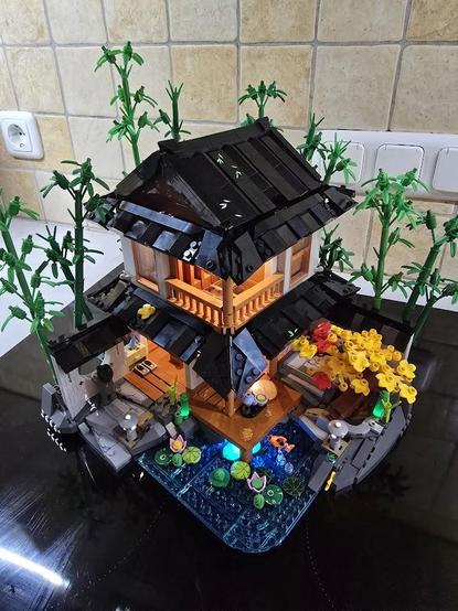 Ein Haus, gebaut aus Klemmbausteinen, in der Art einer Bambushütte, mit einem kleinen Teich im Vorder- und einem kleinen "Bambuswald" im Hintergrund. Ein paar Elemente sind beleuchtet.