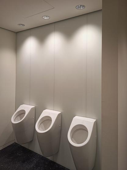 A modern public restroom with three white urinals mounted on a clean, light beige wall. The design is minimalist, with no partitions between the urinals. The lighting is recessed and casts three evenly spaced pools of light above the urinals. The dark flooring contrasts with the smooth, pale walls, emphasizing a sleek and contemporary atmosphere.