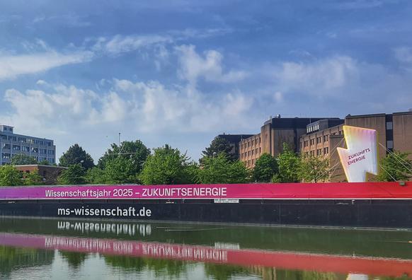 Der mittlere Teil der MS Wissenschaft vor dem Finanzamt Hannover; aufgenommen von der gegenüberliegenden Seite des Mittellandkanals. Die Schriftzüge "Wissenschaftsjahr 2025 - ZUKUNTSENERGIE" und "ms-wissenschaft.de" am Rumpf spiegeln sich im Wasser.
