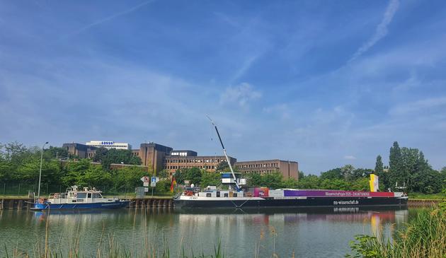 Die MS Wissenschaft in voller Länge von schräg hinten von der gegenüberliegenden Seite des Mittellandkanals aufgenommen. Vor dem Heck liegt ein Boot der Wasserschutzpolizei. Am Ufer sind viele grüne Büsche und Bäume; dahinter das langgestreckte braune Gebäude des Finanzamts; darüber ein blauer bewölkter Himmel.
