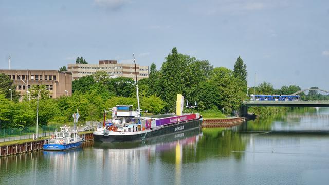 Ein Boot der Wasserschutzpolizei und die MS Wissenschaft links am Ufer des Mittellandkanals; von einer Brücke aus aufgenommen. Am rechten Bildrand eine weitere Kanalbrücke, über die gerade eine blaue Straßenbahn fährt.