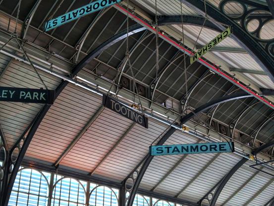 London transport location signage hanging from an arched metalwork roof structure. One sign reads "tooting".