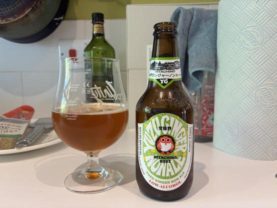 A glass of amber-colored non-alcoholic ginger beer is placed next to a green bottle labeled "Hitachino Yuzu Ginger Non-A". The background features a roll of paper towels, a bottle of olive oil, and kitchen utensils.