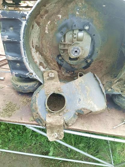 Underside of a lawnmower. The plastic cowl shielding the drive belt is removed. Two hand made metal tangs are bolted to it, along with a previous repair with metal straps and rivets underneath it.