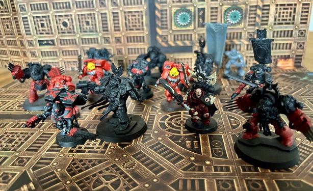 Eine Gruppe von Blood Angels Space Marines steht kampfbereit in einem industriellen Innenraum. Einige Figuren sind vollständig bemalt in Rot und Schwarz, andere noch unbemalt oder grundiert. In der Mitte stehen ein Kommandant mit Kettenschwert und eine ikonische Terminator-Einheit mit Klauen.