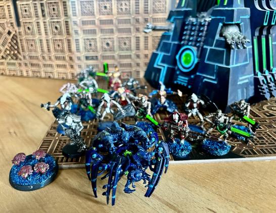 Eine Gruppe von Necron-Miniaturen mit metallisch glänzender Bemalung, grün leuchtenden Waffen und blauen Basen steht vor einer futuristischen Pyramidenstruktur mit leuchtenden Neonkanten. Im Vordergrund sind auch Drohnen und ein skorpionartiges Kriegsgerät zu sehen.