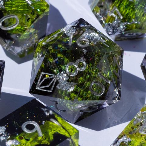 A 20-sided die in the middle, surrounded by other TTRPG dice. The die contains green snake and dark glittery swirls and is inked in silver.