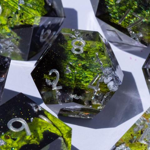 A 12-sided die in the middle, surrounded by other TTRPG dice. The die contains green snake and dark glittery swirls and is inked in silver.
