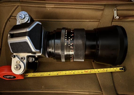 This is the long lens, on the Pentacon Six medium format camera body. The lens hood makes it even longer. It sits on a brown suitcase and a yellow metal  retractable ruler beside shows almost a foot long.