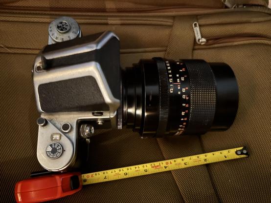 This is the long lens without the hood. The large medium format Pentacon Six camera and SLR viewfinder are seen on a brown suitcase. The yellow ruler shows several inches long.