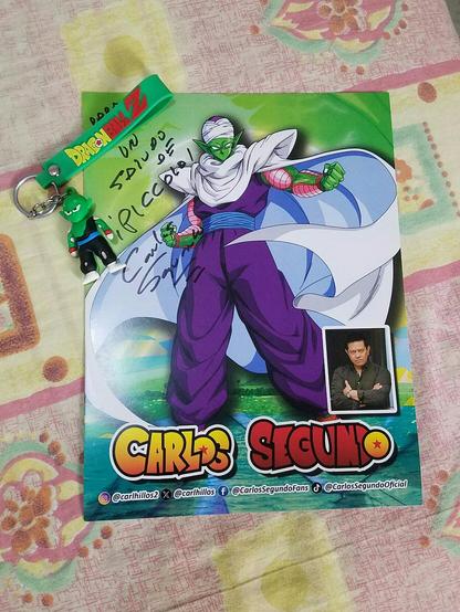 Foto ofrecida por Carlos Segundo en su mesa de firmas, donde vemos al personaje de Piccolo, el nombre del invitado y su foto personal en un pequeño recuadro en la parte inferior, junto a sus redes sociales. Vemos en la esquina izquierda superior la firma personalizada  y tapando mi nombre un llavero de Piccolo que llevé ese día en mi bolso.