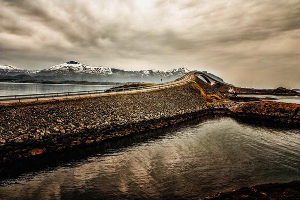 A winding bridge stretches across rocky terrain leading to snow-capped mountains in the background. The scene features muted colors, a calm water surface, and a cloudy sky, suggesting a serene and remote landscape. Norway, Altanterhavsvegen, 2016.