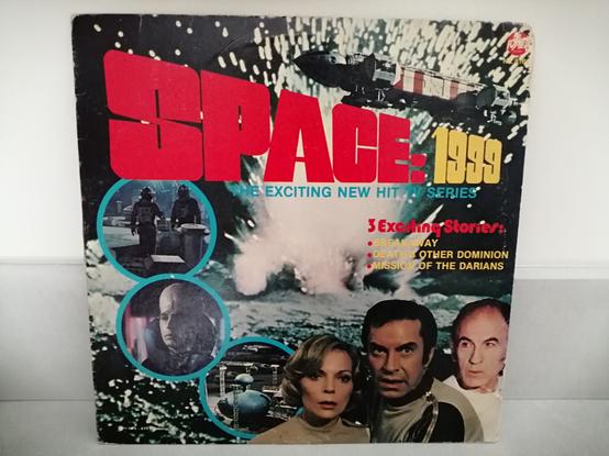 Sur un fond noir une explosion envoyant des étincelles. Au dessus un vaisseau spatial "Eagle". En bas le visage de 3 acteurs de la série : Barbara Bain, Martin Landau et Barry Morse. En haut et en gros le tire SPACE en rouge et 1999 en jaune.