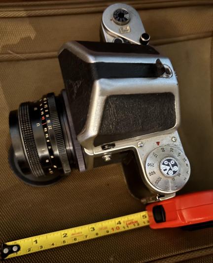 The large, medium format Pentacon Six camera and SLR viewfinder (they’re removable) is seen with the standard lens, in this case 80mm focal length. A yellow retractable ruler shows several inches. The camera sits on a brown suitcase.