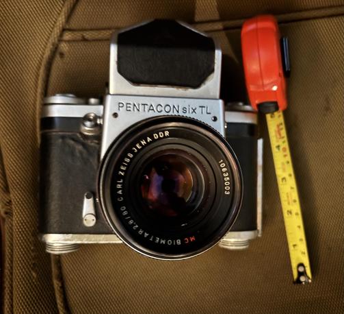 The 80mm Zeiss lens is seen facing the camera. The face of the large, medium format Pentacon Six is seen in front of the same brown suitcase. The name is seen, “Pentacon six TL” (for through the lens).
