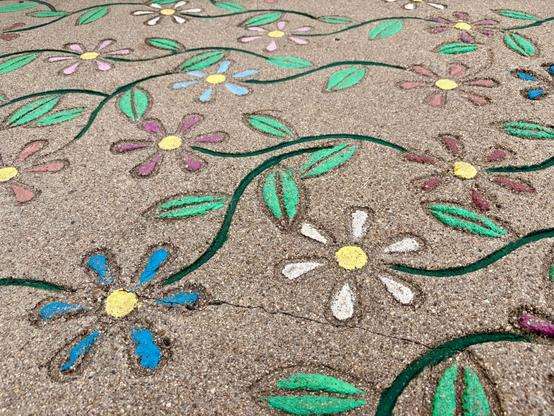 Painted colorful flower designs on a concrete surface. Pink, blue, white, and yellow flowers with green leaves connect via curving vines. The flowers have simple daisy-like designs with round centers and surrounding petals. The chalk creates a vibrant garden pattern against the gray cement background.​​​​​​​​​​​​​​​​