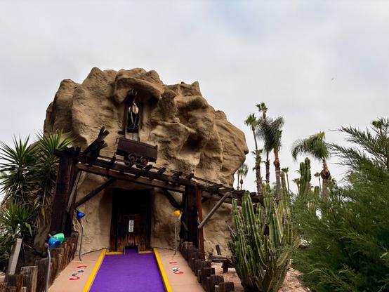 This miniature golf course hole features a large artificial rock formation designed to look like a mine. A purple carpet path leads to a wooden door at the base. A small mining cart sits on wooden tracks above the entrance. The scene includes desert landscaping with palm trees, cacti, and other plants surrounding the rock structure. Colorful lights decorate the entrance path.​​​​​​​​​​​​​​​​