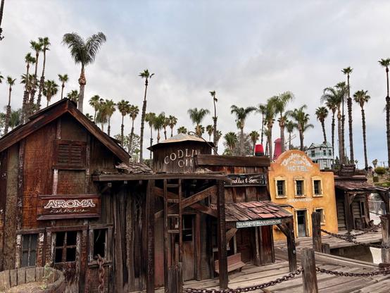 The image shows an old Western-style miniature town with weathered wooden buildings. The most prominent structures include an "AROMA Fish Packers" building, "CODLIVER OIL" tank, and a yellow "HOTEL" building. Several business signs are visible including "Marine Hardware" and another barely legible sign. Palm trees tower above the scene against a cloudy sky. Chain barriers separate visitors from the display.