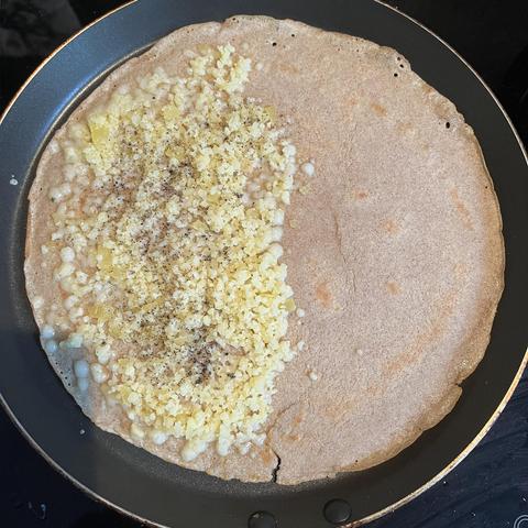 Ein Galette in einer Galettepfanne auf einem Herd, mit geriebenem Käse bedeckt.