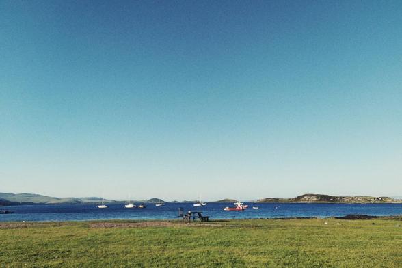 A vertical colour photograph of a field of grass by an inlet. In the distance, low hills mark the horizons. Boats float on the water. The sky is bright summer blue.