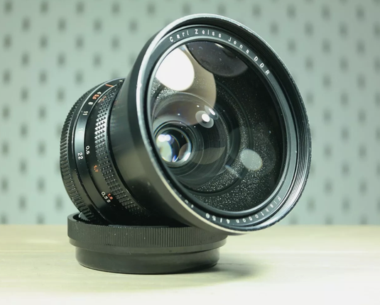 A large camera lens is sitting on its lens cap at an angle. The lens reads in part "Carl Zeiss Jena DDR". The table is a pale wood and the background is out of focus dotted wallpaper, pale green.