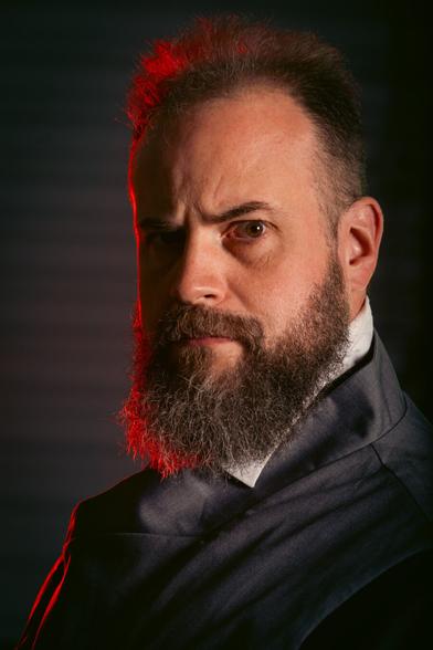 This is a dramatic portrait of a bearded man with a stern expression, lit with intense contrast and a striking red accent light on the side of his head and beard. He is wearing a dark, high-collared garment .