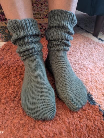 Picture of two human feet, visible from mid-shin down, wearing hand knit, slouch socks in in a slate gray/green color. Cuffs are ribbed in a knit one/purl one, ribbing pattern. Socked feet are resting on a patterned, shag carpet with burgundy and pink colors underfoot. A patterned, multicolored carpeted stool, and base of a teal green couch with one wooden leg is visible in the background.