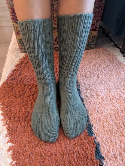Picture of two human feet, visible from mid-shin down, wearing hand knit socks in in a slate gray/green color. Cuffs are ribbed in a knit one/purl one, ribbing pattern and pulled up straight to mid-shin length. Socked feet are resting on a patterned, shag carpet with burgundy, pink, and cream colors underfoot. A patterned, multicolored carpeted stool, and base of a teal green couch with one wooden leg is visible in the background.