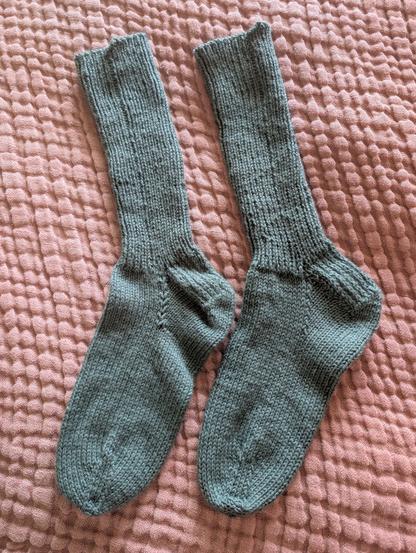 Picture of a pair of hand-knit socks in in a slate gray/green color. Cuffs are ribbed in a knit one/purl one, ribbing pattern. Socks are resting on a terracotta-colored, textured linen blanket.