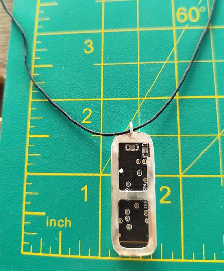 the back side of a silver pendant with an old PCB. it has a black cord and it's placed in green mat