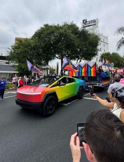 A photograph, depicting a cyber truck covered in a rainbow colored wrap, pulling a float in a gay pride parade.