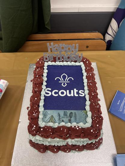 A cake! It is rectangular and in portrait.
Around the edge is a band of red icing, then light blue. Then more red.
On the top is the Sdours logo and the word “Scouts” followed by a print of our 80th anniversary badge.