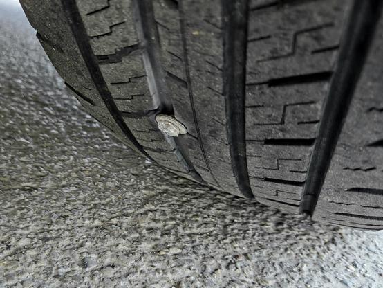 Close-up of a car tire with a silver screw embedded in the tread, captured from a low angle on a rough asphalt surface. The screw is lodged in a groove of the tire, indicating potential risk for a slow air leak or future flat.