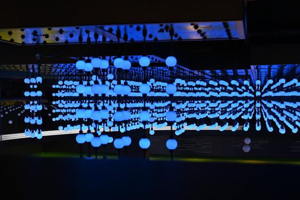 A visually striking installation featuring numerous glowing blue spheres suspended in a grid-like pattern. The lights appear to extend infinitely due to mirrored reflections on the ceiling, creating a futuristic, immersive effect in a dark room.