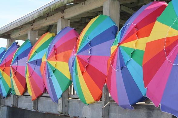 Several rainbow-coloured beach umbrellas in a row decorate the outside of an ocean pier.