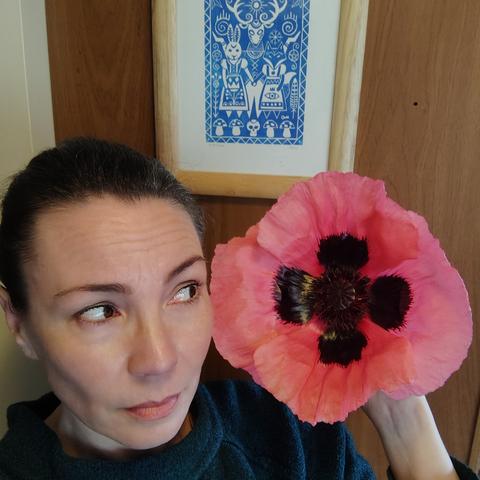 A woman's face next to a pink poppy flower that is roughly the same size as her head.