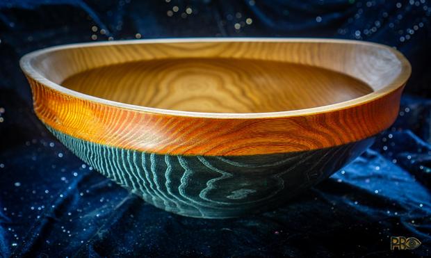 Color photo. Close-up of a large turned wooden bowl, 36 cm diameter 14 cm deep – seen close up from the side, but slightly from above so that you can also see in and down into the bowl. The inside is dark brown elm with clear growth rings. Inlaid with food-safe beeswax oil. The top wide part is stained red, with the growth rings highlighted in gold. The lower part, the body of the bowl, is dark, dark blue almost black, with the growth rings highlighted in silver. The background is a blue tablecloth with glitter.

Fargefoto. Nærbilde av en stor dreid trebolle, 36 cm diameter 14 cm dyp – sett tett inntil fra siden, men litt ovenfra slik at man også ser inn og ned i bollen. Innvendig er den mørk brun alm med tydelige årringer. Innsatt med matsikker bivoksolje. Utveding er øverste brede parti beiset rød, med årringene uthevet i gull. Nedre del, bollens kropp, er mørk, mørk blå nesten svart, med årringene uthevet i sølv. Bakgrunnen er en blå duk med glitter.