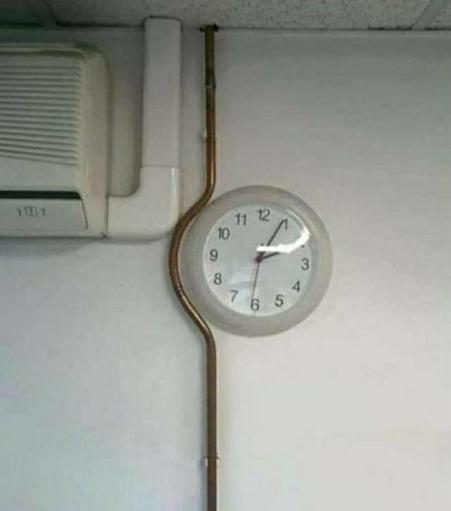 A photograph of a clock on a wall. A copper plumbing pipe descends vertically, but is curved by whoever installed it to curve around the clock