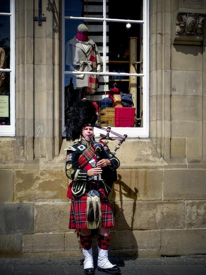 A man in traditional Scottish regalia, including a black feathered hat, detailed jacket, and red tartan kilt, plays bagpipes on a sidewalk. Behind him, a store window displays various items like scarves and a mannequin dressed in a beige coat. The building's architecture features distinct stone work and tall, rectangular windows.