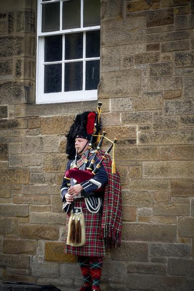 A bagpiper in traditional Scottish attire stands against a stone wall next to a white-framed window. He wears a black feather bonnet, a tartan kilt with a matching plaid draped over his shoulder, and a dark doublet with decorative silver buttons and badges. He plays a set of bagpipes, featuring prominent drone pipes and a red and black fabric cover. His look is completed with red and green tartan hose and black shoes, while a sporran hangs at his waist.