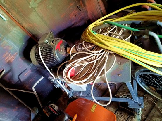 Back of a darkly shed. Under a hose and a long CEE 3-phase cable, a wheeld table on which a very big electric motor is mounted. On the shaft of the motor, a screw threaded cone for splitting logs by screwing into them until they pop. There's an electric junction box under the table. A big red arrow painted on the front of the motor indicates direction of rotation.