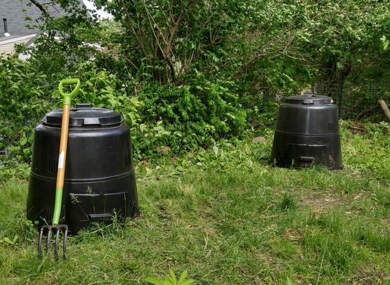 A backyard with two black composter bins, with a green handled garden fork next to one of them. In the background are lots of trees and bushes.
