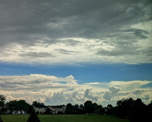 A landscape photo with puffy, turbulent clouds beyond a row of houses and trees. In the foreground is a green field and a layer of darker gray clouds is directly overhead. In between the distant and nearby clouds is blue sky.