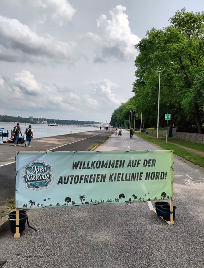 Ein Photo der für Menschen geöffneten Kiellinie Nord. Im Vordergrund ein Banner mit der Aufschrift: Willkommen auf der autofreien Kiellinie Nord!