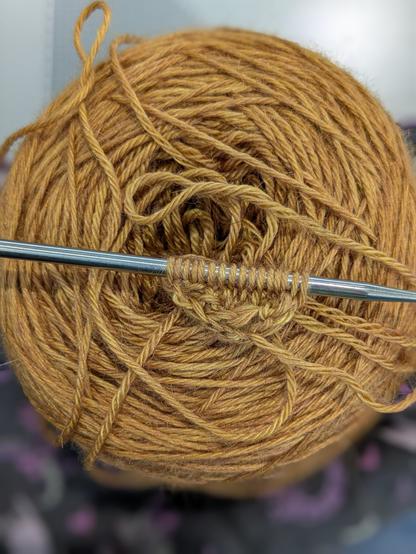 A cake of golden yarn. A steel knitting needle is sitting on top with the tiniest (two rows) work in progress
