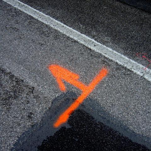 Square section of an asphalt surface with a fine white stripe extending from the top left to the center of the picture's right edge. Below the line, an arrow with a crossbar at its bottom has been painted in bright orange. The picture is dominated by the contrast of orange and white. The asphalt is also not uniformly dark. At the picture's lower edge, a triangle is darker, a recently repaired piece of asphalt.