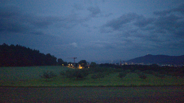 Blick während der Blauen Stunde über Wiesen und Felder auf das Westende der Siedlung Rütihof mit einigen natriumdamp-gelben Strassenlaternen. Rechts im Hintergrund der Uetliberg. Der Himmel ist blau mit ein paar Wolken in diversen Blautönen.