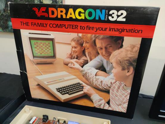 A Dragon 32 retail box, with a photo of models posing as the prototypical white middle class family at whom the computer was targeted. In a row, from furthest from the camera to closest, we see a little brown-haired girl who is looking at the computer screen and smiling. She's wearing a tan blouse. Her hands are folded in front of her, elbows on the table. Next to her is mum, with strawberry blonde hair, also smiling and looking at the screen. It's not possible to determine her hair length; she appears to be wearing a cream coloured blouse. Mum has her arm around the little girl's shoulder. Next to her is dad, fortyish, with an executive haircut of the era, smiling and looking at the son's eyeline. Dad is wearing a white shirt open at the collar. In the foreground is son, very blond, wearing a comfy cream sweatshirt with double bands of rusty stripes. He is looking at the screen. His left hand is on the Dragon computer keyboard and his right hand points to the screen. The computer is placed on a wood veneer table; the edge of some framed art is visible on the magnolia wall.

The word "DRAGON" has the colour spectrum reversed out of it, with the dragon logo to the left at the red end of the spectrum. The text "The family computer to fire your imagination" appears underneath.